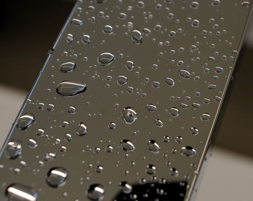 Close-up of a stainless steel surface, likely a kitchen countertop or appliance, featuring numerous water droplets scattered across its smooth, reflective, metallic finish. The surface appears to be r