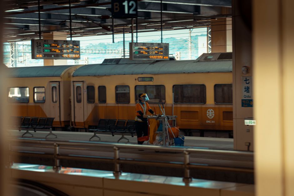 A woman with dark skin and short black hair is standing in a modern kitchen area, holding a vacuum cleaner with yellow and black components. She is dressed in a grey T-shirt and bright orange overalls